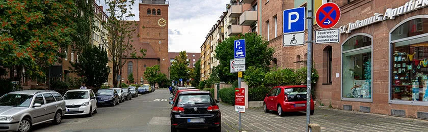 Wohngebiet mit mehreren parkenden Fahrzeugen. Ein rotes Fahrzeug parkt unerlaubt vor einer Apotheke auf dem Gehweg. Parkverstoß